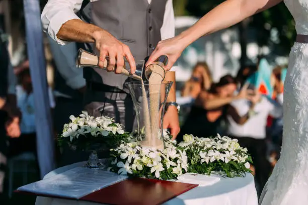 The bride and groom are pouring sand together at a wedding ceremony at a hotel. On the table was a beautiful bouquet of white flowers. There were many guests in attending