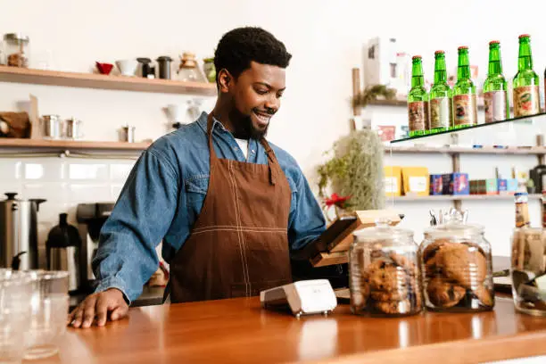 Black bearded man wearing apron working with cash register in cafe indoors