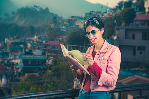 In this outdoor dusktime image, an Asian/Indian beautiful young woman reads a book happily, standing by the railing on the rooftop of her house in the hills of Himachal Pradesh. She is in a pink denim jacket, blue jeans, and tinted sunglasses.