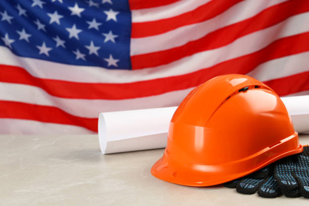 istock Orange protective hard hat, gloves, construction drawing on light grey table against American flag. Space for text 1454205653