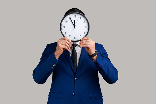 Unknown anonymous man hiding face behind big wall clock, afraid of deadline, wasting time, being late, wearing official style suit. Indoor studio shot isolated on gray background.