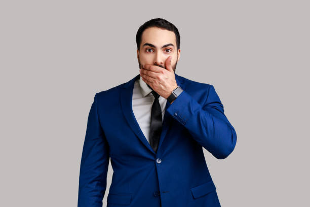 Attractive bearded businessman closing mouth with hand, afraid to say too much, embarrassment. Portrait of attractive bearded businessman closing mouth with hand, afraid to say too much, embarrassment, wearing official style suit. Indoor studio shot isolated on gray background. closed mouth stock pictures, royalty-free photos & images