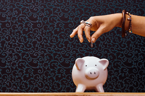 Young woman's hand, adorned with plentiful costume jewelry, about to insert a coin into a china piggy bank.