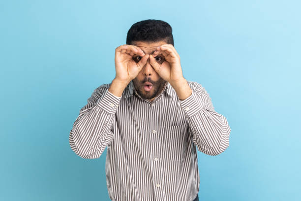 istock Amazed young businessman looking through fingers shaped like binoculars and expressing excitement. 1454209478