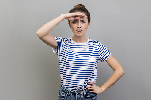 Portrait of beautiful ambitious woman wearing striped T-shirt watching far away with hand above eyes, looking forward to future. Indoor studio shot isolated on gray background.