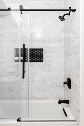 A remodeled glass shower with marble subway tiles, a shelf with black hexagon tiles, and a sliding glass door with black hardware.