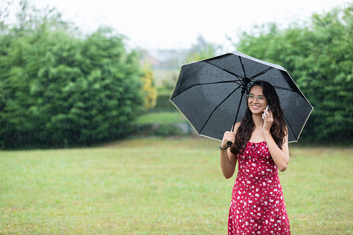 Cheerful young Hispanic female millennial with long dark wavy hair in summer dress and eyeglasses smiling and looking away while talking on smartphone standing in park with umbrella on rainy day