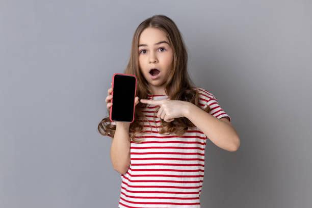 istock Little girl pointing finger at smartphone with white empty display, shocked by application. 1454211692
