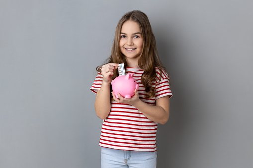 Portrait of smiling adorable little girl wearing striped T-shirt standing holding putting banknote into pink piggy bank in hands, saving money. Indoor studio shot isolated on gray background.