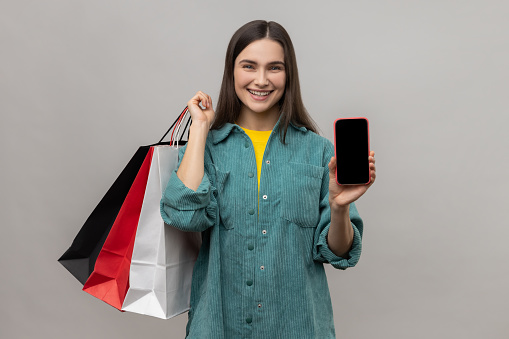 Happy woman standing with satisfied look showing shopping bags and smart phone with blank screen for advertisement, wearing casual style jacket. Indoor studio shot isolated on gray background.
