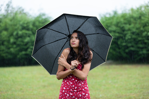 Upset young ethnic freezing female in summer dress with umbrella rubbing arms and looking up on grassy lawn in park on rainy day