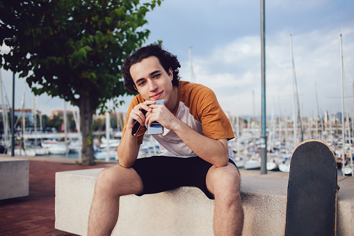 Cute curly haired guy in casual wear resting on rocky fence smiling and looking at camera while leaning elbow on knees on sunny day in Barcelona