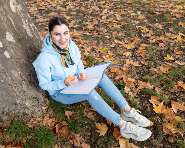 eine studentin sitzt mit einer blauen mappe in den händen unter einem baum und lächelt, österreich, salzburg - fall semester stock-fotos und bilder