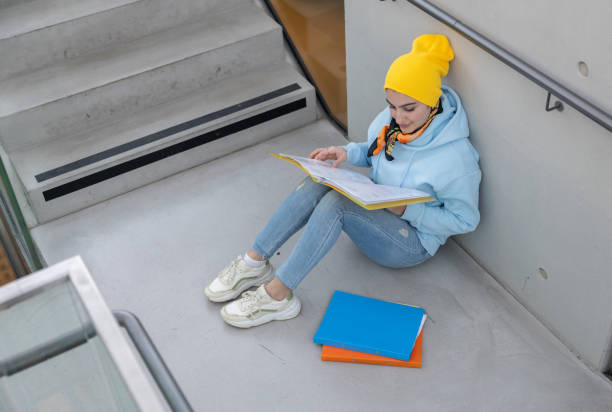 eine studentin in blauem sweatshirt und gelber mütze sitzt auf einem steinboden zwischen treppen und wartet auf den unterricht - fall semester stock-fotos und bilder