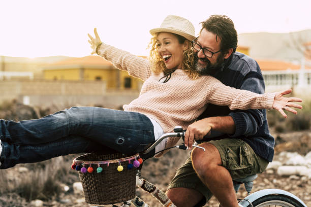 Happy couple enjoy outdoor leisure activity together carrying and using a bike and laughing a lot. Love and friendship with mature man and woman in youthful lifestyle. Concept of joyful and excitement stock photo
