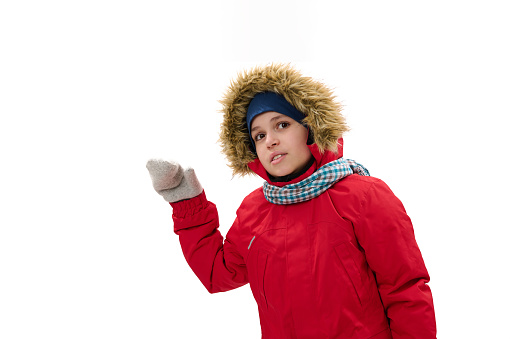 Adorable Middle-Eastern teenage boy, in red down parka with fur hood, blue scarf and woolen mittens, smiles looking at camera, points at copy space for your ads, white background. Cool weather. Winter