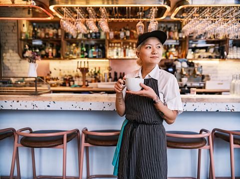 Young Korean female chef having morning coffee at work. She is wearing usual kitchen attire with apron and baseball cap. Interior of a busy restaurant.