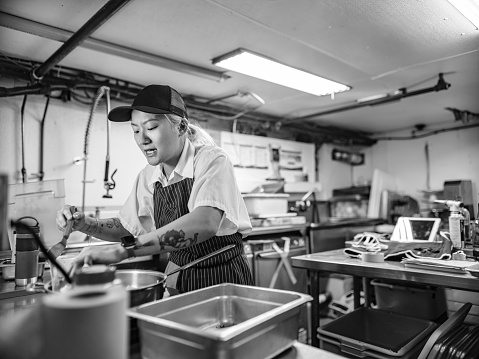 Young Korean female chef working in restaurant kitchen. She is wearing usual kitchen attire with apron and baseball cap. Interior of industrial kitchen of a busy restaurant.