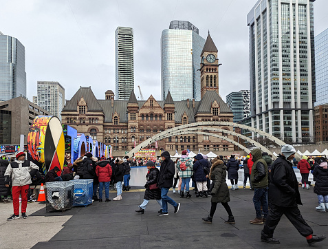 Toronto, Canada - December 10, 2022: Festival goers attend Holiday Fair in the Square at Nathan Phillips Square, City Hall. This celebration includes a midway, Christmas market and entertainment. The fountain in front of the Indigenous medicine wheel and Toronto Sign converts to an ice rink under the arches. The Old City Hall with the clock tower stands on Queen Street West in the background.