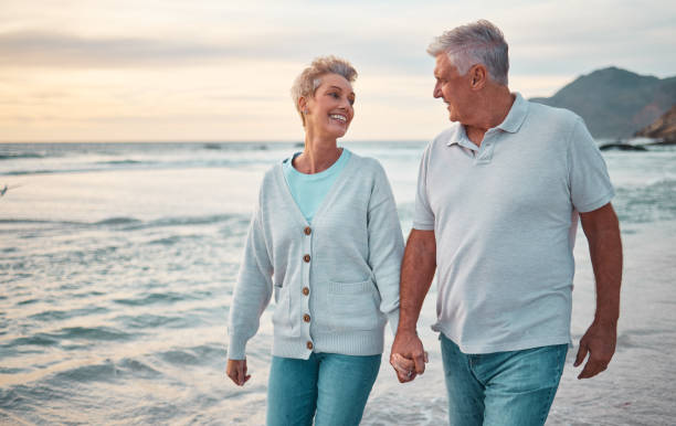 Beach, holding hands and senior couple with sunset for outdoor wellness, retirement holiday and outdoor date mock up sky. Happy elderly people, or old man and old woman walking by sea or ocean water playa, tomados de la mano y pareja mayor con puesta de sol para el bienestar al aire libre, vacaciones de jubilación y fecha al aire libre simular el cielo. ancianos felices, o ancianos y ancianas caminando por el mar o el agua del océano