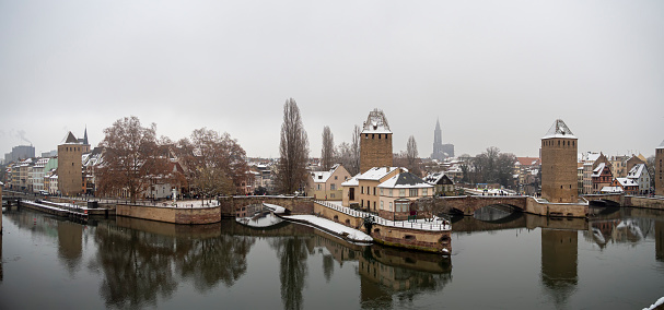 Panoramic of La Petite France a part of Strasbourg city during winter. Snowy houses reflect on river that splits into canals. Panorama of medieval bridges Ponts Couverts and Cathedral.