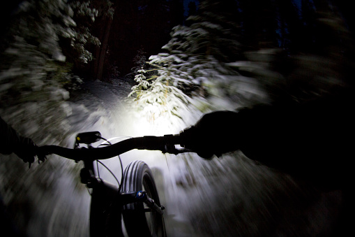 The handlebar view of a man going for a winter fat bike night ride. Fatbikes are mountain bikes with oversized wheels and tires for riding on the snow. His headlight is mounted on his handlebar and lights the trail in front of him.