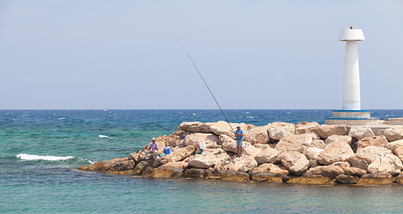 Ayia Napa, Cyprus - June 12, 2018: Fishermen are on breakwater near white lighthouse tower, Agia Napa port