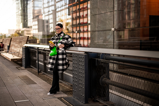 Fashionably dressed woman with eyeglasses wearing black and white jacket and a green bag seen standing by the fence on the street and texting on her phone in Manhattan, New York, during a coffee break.