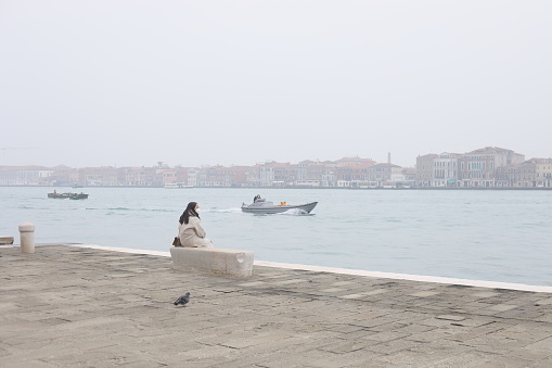 Japanese tourist wears a surgical mask  while sitting alone  on  a bench in the Fondamenta Zattere in Venice - Italy during the Covid 19 pandemic. In the background the Giudecca canal and two motor boats with workers