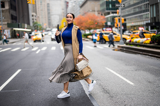 Beautiful woman in a beige sleeveless coat, skirt, turtleneck sweater, white sneakers and with a handbag seen running across the street in Manhattan, New York, while using her wireless headphones and talking on the phone during a break at work.