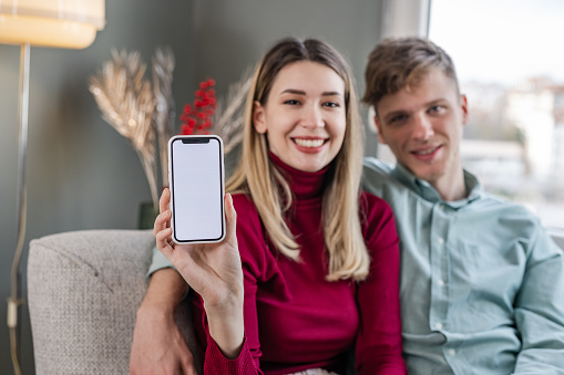 Cheerful young couple sitting in the living room, smiling and showing smartphone with white screen to the camera. Chroma Key. Copy space.