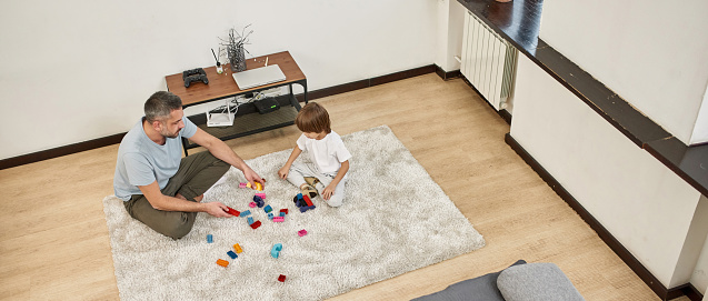Top view of father and little son playing with construction building blocks on carpet at home. Young caucasian family relationship. Fatherhood and parenting. Domestic leisure, hobby and entertainment