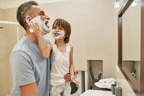 Little boy smearing shaving foam on father beard at home bathroom. Cheerful caucasian child and man joking and spending time together. Dad looking at mirror. Fatherhood. Face skin care and hygiene