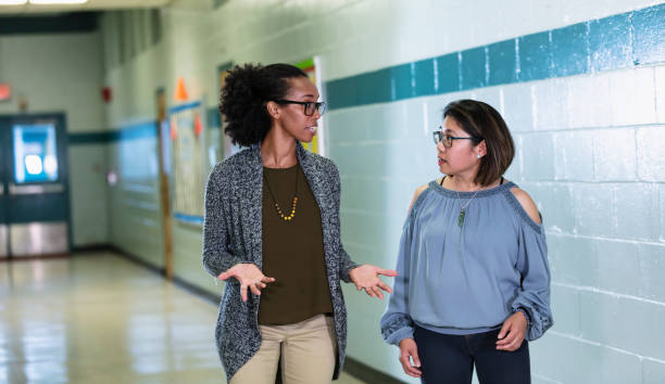 Multiracial teachers converse, walking in school hallway stock photo