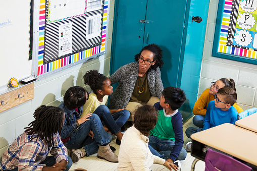 A multiracial group of seven school children and their teacher doing a practice drill, sheltering in place, sitting on the floor in a corner of their classroom.