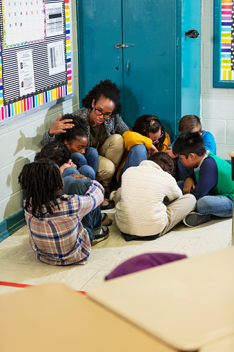 A multiracial group of seven school children and their teacher doing a practice drill, sheltering in place, sitting on the floor in a corner of their classroom.