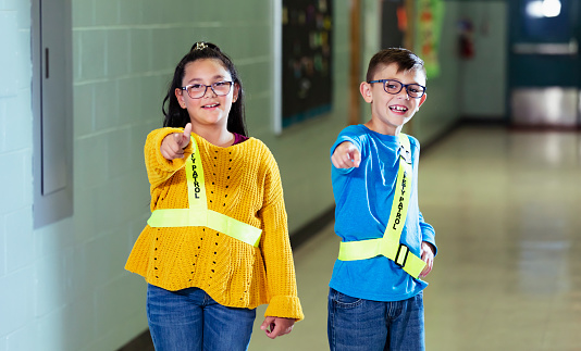 Two multiracial elementary students, proud members of the school safety patrol, standing in the school hallway, smiling and pointing at the camera. The children are 9 years old. The girl is Hispanic.