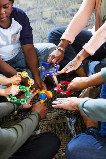 Cropped, high angle view of a group of three multiracial children and two mothers sitting together on the floor in a school library making music with toy tambourines and maracas. The women are in their 30s and the children are 8 to 11 years old.