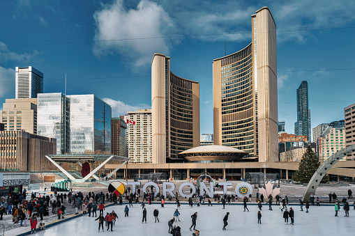 People skate on the Nathan Phillips Square skating rink in downtown Toronto, Ontario, Canada on a sunny day, with the City Hall building in the background.