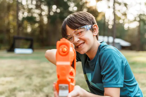A tween boy wearing protective eyewear smiles and takes aim while playing with a nerf toy gun outside in the backyard of his home on a warm and sunny afternoon.