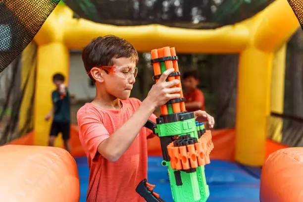 A boy loads foam ammunition in a toy nerf gun while having a fun battle with his friends in an inflatable bouncy house during a backyard birthday party.