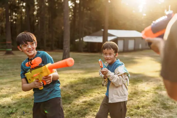 Two multiracial brothers defend each other while having a nerf gun battle with their dad outside in the back yard on a warm and sunny afternoon. Both boys are wearing protective eyewear.