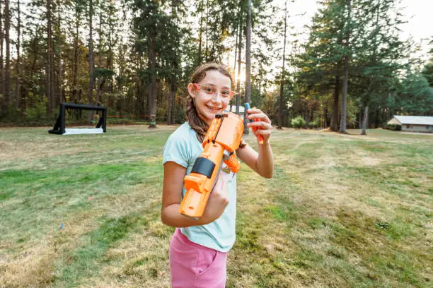 Medium range shot of a tween girl smiling directly at the camera and putting foam ammunition in a toy gun while playing outside in the back yard of her rural home on a warm evening.