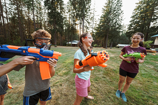 A multiracial group of boys and girls stand outside in the grass and laugh while having a playful battle with toy nerf guns. The kids are wearing protective eyewear.