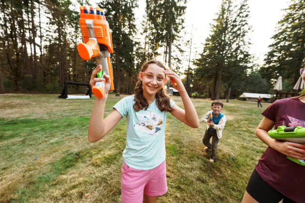 A tween girl wearing protective eyewear smiles directly at the camera and holds a toy nerf gun in while playing outside in the back yard with her cousins.