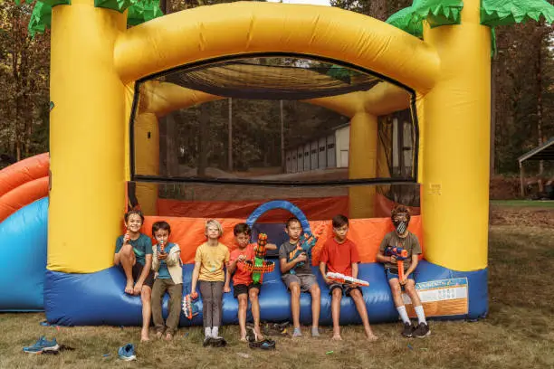 A multiracial group of elementary age and tween boys sit in a row along the edge of an inflatable bouncy castle and hold toy nerf guns while at a back yard birthday party.