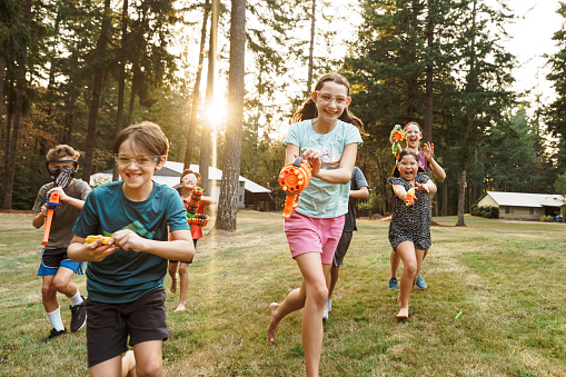 Medium range shot of a multiracial group of boys and girls running in the grass and aiming toy guns at the camera while having a playful and fun nerf gun battle at an outdoor birthday party. In the background the setting sun shines through a forest of fir trees.