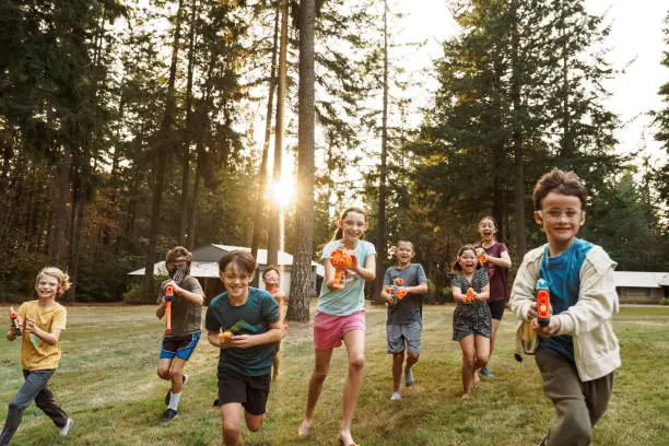 Medium range shot of a multiracial group of boys and girls running in the grass and aiming toy guns at the camera while having a playful and fun nerf gun battle at an outdoor birthday party. In the background the setting sun shines through a forest of fir trees.