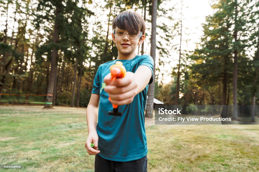 Tween boy aiming toy gun toward camera A Eurasian tween boy wearing protective eyewear playfully points a small toy nerf gun toward the camera while playing outside in the back yard of his rural home. 12-13 Years Stock Photo