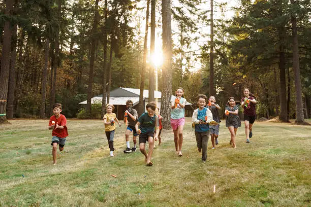 An excited group of elementary age and tween boys and girls run toward the camera while having a playful nerf gun battle outside in a large rural back yard during a fun birthday party. In the background the setting sun shines through a forest of fir trees.